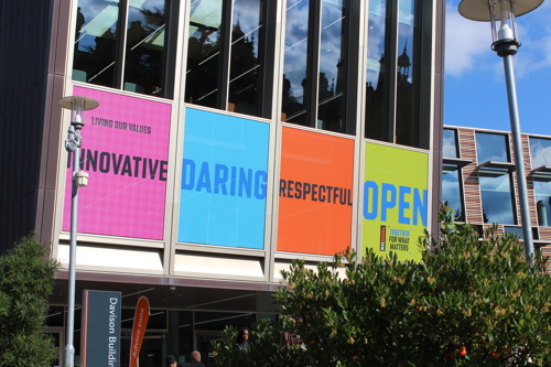 A photograph showing a close up of the Emily Wilding Davison Building on campus, Across the bottom of the windows are brightly coloured posters highlighting the words of the new RH2030s Values - Innovative, Daring, Respectful, Open
