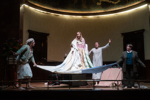 A picture of four people on a stage mid-performance. A person in a long white costume seems to be standing on a large sheet held by the other three performers.